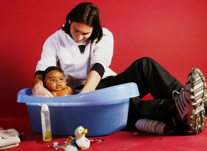 A surreal studio photograph against a red backdrop showing a man in athletic wear giving a baby a bath inside a small blue plastic tub on the floor.