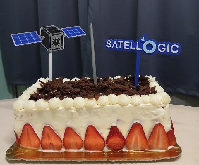 A white frosted cake decorated with strawberries around the base and chocolate shavings on top, featuring two toppers: a satellite illustration and a 'SATELLOGIC' logo.