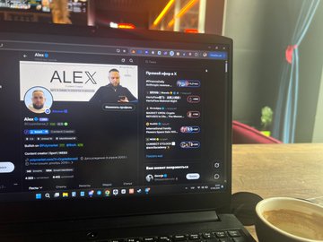 A photograph of an open laptop on a table displaying a live stream featuring a person named 'ALEX' with Russian-language chat, alongside a cup of coffee in the foreground.