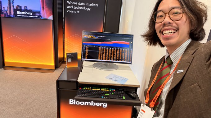 A smiling man with glasses wearing a blazer and tie standing next to a Bloomberg-branded trading station with dual monitors and an orange accent keyboard.