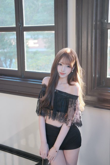 A young woman with long brown hair poses near a window, wearing a black off-shoulder lace top and matching shorts, exuding elegance that supports the post's question about top-tier beauty.