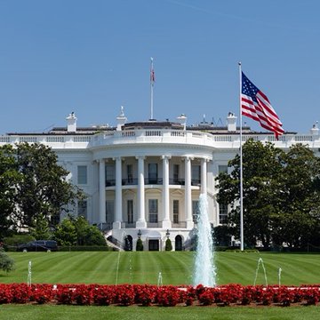 The White House is shown from the South Lawn, with the American flag prominently displayed against a clear blue sky and a fountain spraying water over red flower beds. The image visually anchors the accompanying post about the White House's stance on AI regulation, reinforcing the official government setting where Kevin Hassett's comments were made regarding private-sector-led approaches to AI safety.