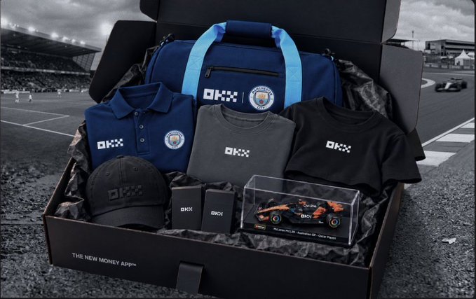 A promotional gift box from OKX titled 'THE NEW MONEY APP™' containing Manchester City-branded merchandise including a blue duffel bag, polo shirt, t-shirts, cap, and a model Formula 1 race car.