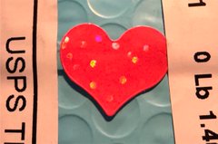 A hand-painted enamel pin featuring a red heart with colorful sprinkles rests on a textured surface between two USPS postage labels, celebrating the Trencher Sol prerelease exclusive TCG drop on dyli.io signed by artist @grizzle_art.
