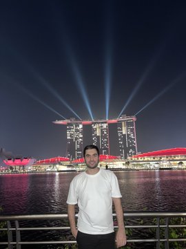 A man in a white t-shirt stands in the foreground at night with the illuminated Marina Bay Sands hotel in Singapore visible behind him across the water, featuring its iconic light show.