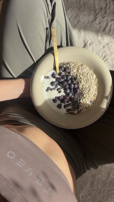 A person holds a bowl containing oatmeal, blueberries, and chia seeds with a wooden spoon, sitting on a bed with 'OEWE' visible on their shorts.