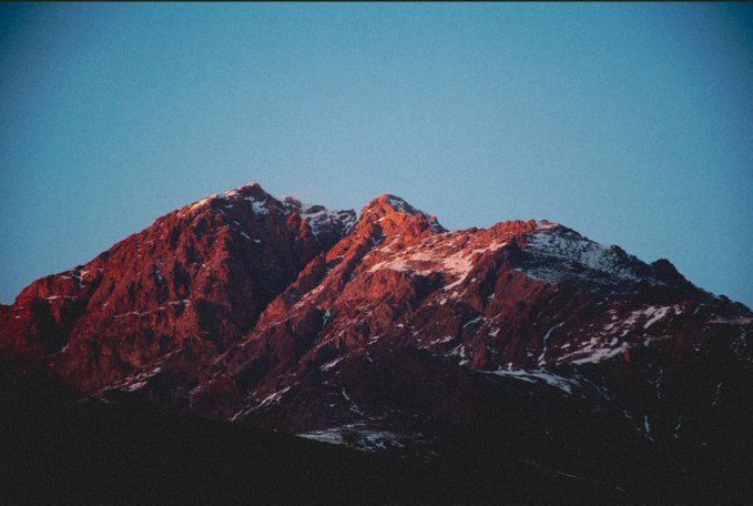 A photograph of a mountain peak illuminated by reddish alpenglow against a deep blue twilight sky, with patches of snow visible on the dark rocky slopes.