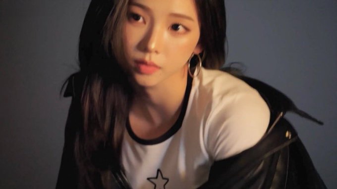 A young woman with long dark hair, wearing a white ringer t-shirt with a star and a black jacket draped off her shoulder, looks at the camera in a softly lit portrait.