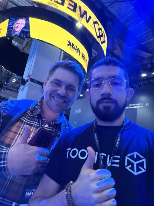 Two smiling men pose for a selfie at the TOKEN2049 event, with one wearing a "ROAM" lanyard and the other in a "100MOVE" branded shirt, standing beneath a vibrant yellow "ROAM" display arch as they invite attendees to find them at booth PB1-21.