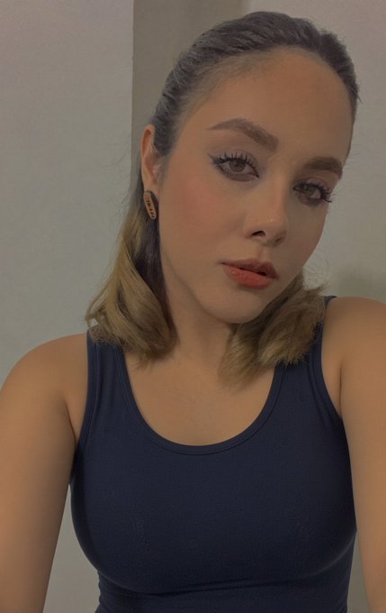 A young woman with shoulder-length brown hair poses for a selfie, wearing a dark navy tank top with makeup and a gold hoop earring.