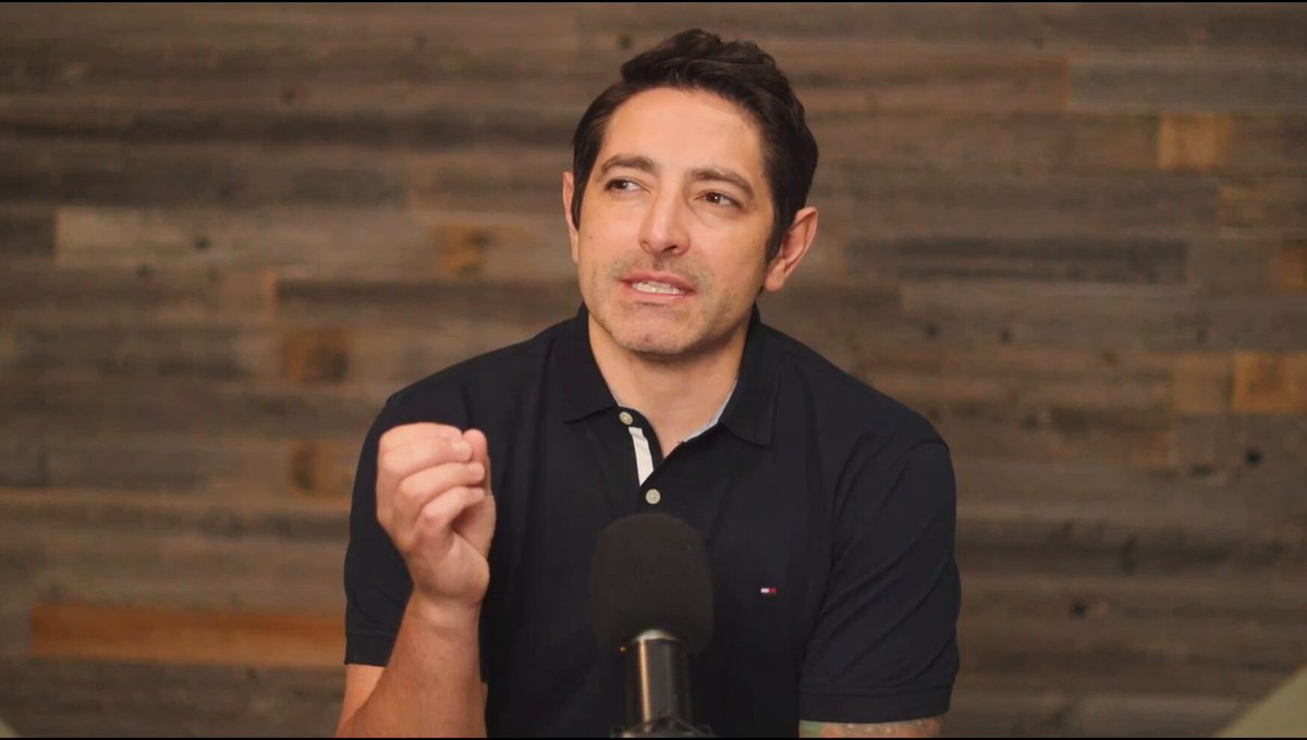 The image shows a man wearing a black polo shirt, sitting at a microphone and speaking into it. The background features a wooden wall or paneling, which adds an element of rustic charm to the scene. The caption provided seems unrelated to the main subject matter but includes some technical terms like "sub-transaction privacy", "deterministic finality", and "conditionally executable contracts".