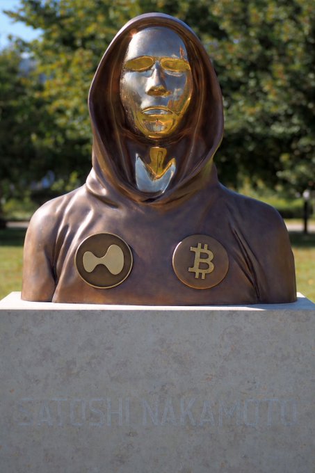A bronze statue of a hooded figure with a golden face, featuring XRP and Bitcoin emblems on the chest, mounted on a pedestal inscribed with 'SATOSHI NAKAMOTO'.