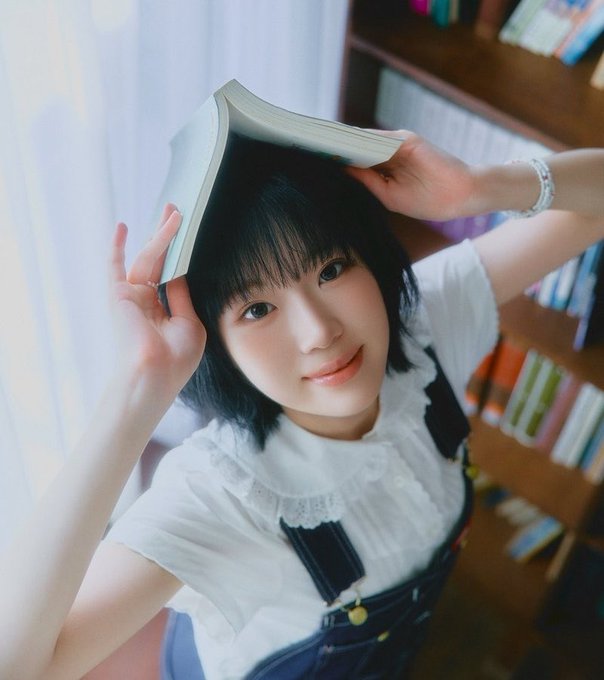 A young woman with short black hair holding an open book over her head like a hat, wearing a white blouse with lace details and denim overalls, standing near a bookshelf in soft lighting.