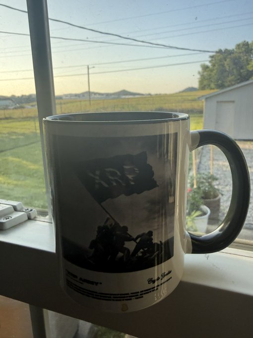 A white coffee mug displaying the iconic Iwo Jima flag-raising image with "MARINE" text and "Victory at Iwo Jima" inscribed, sitting on a windowsill overlooking a sunny vineyard landscape.