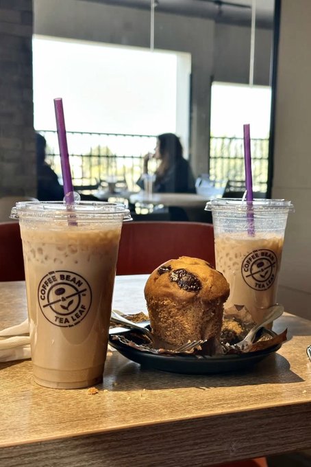 The image shows a wooden table with two cups of coffee and a muffin placed on top. The coffee cup has a straw sticking out of it, while the other cup does not have one. There are also spoons visible near the coffee cups. In the background, there is another person sitting at a table in what appears to be a cafe or restaurant setting.