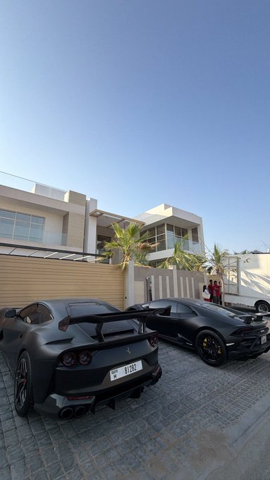 Two black luxury sports cars, including a Ferrari with license plate '81292', are parked in the driveway of a modern villa.