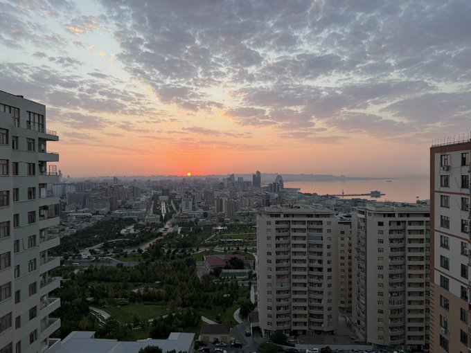 A sunset view over a coastal city with apartment buildings, a park, and water visible in the distance.