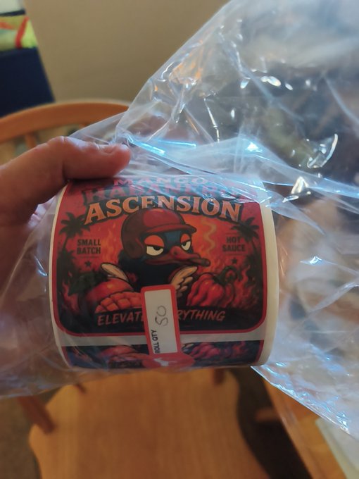 A hand holding a 'Rising Ascension' hot sauce bottle (small batch, QTY 50, 'Elevate Everything') wrapped in a plastic bag, featuring a cartoon duck in a red cap on the label.