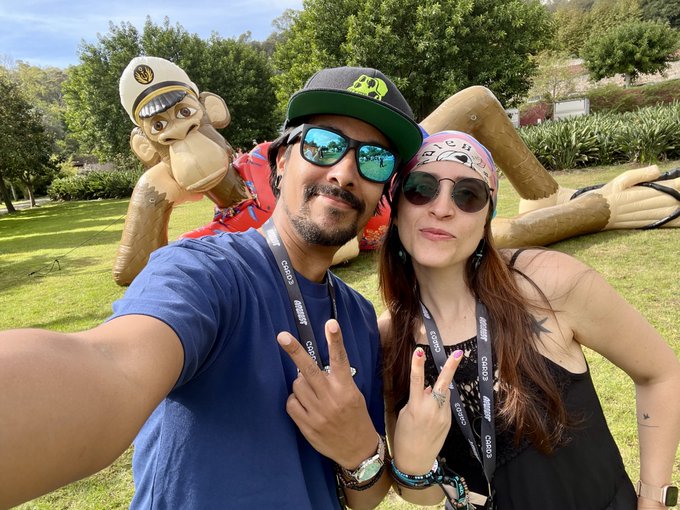 A couple takes a selfie in a grassy park while throwing up peace signs, posing in front of a large inflatable monkey statue wearing a captain's hat and red shirt, fitting the "GM Apes" vibe. Both are wearing lanyards with visible "CASIO" branding and "CHIPS" text, along with stickers featuring ape-related imagery tied to the NFT community.