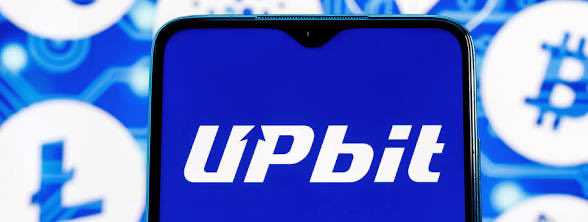 Close-up of a smartphone displaying the "UpBit" logo on a blue screen, with cryptocurrency icons (including Litecoin and Bitcoin) in the blurred background.