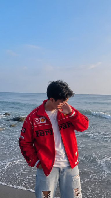 A person wearing a red Ferrari jacket and a white shirt, standing on a beach with the ocean in the background, with their hand partially covering their face.