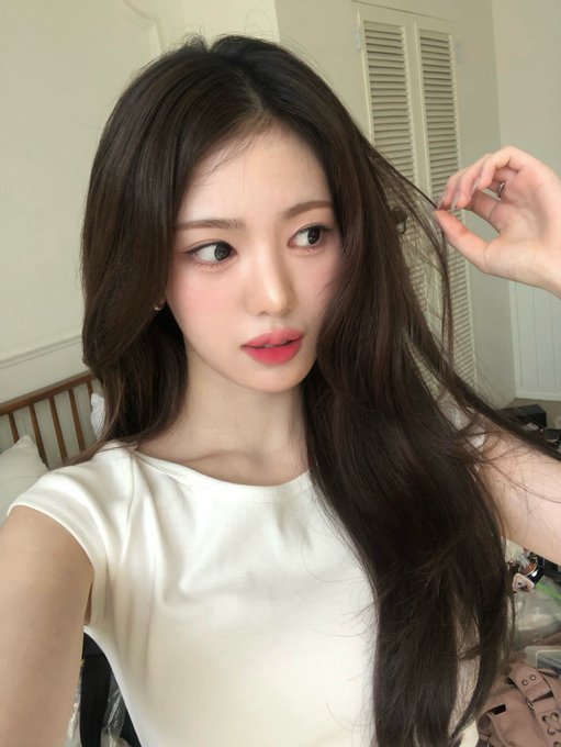A young woman with long dark hair and red lipstick takes a selfie, wearing a white t-shirt in a room with white paneled walls.