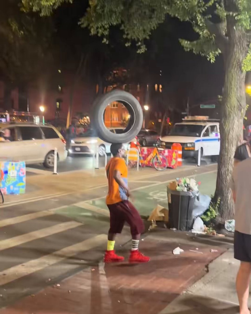 The image shows a man walking down a sidewalk, balancing a tire on his head while wearing an orange shirt and red shoes. In the background, there are several cars parked along the street, including one car that appears to be a police car. The scene also includes a traffic light in the distance.