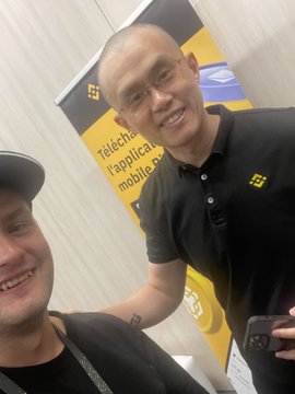Two men smile for a selfie at what appears to be a crypto event, standing in front of a Binance promotional banner with text referencing "l'applicazione mobile" (Italian for "the mobile application"), visually confirming the casual encounter with Binance founder CZ (Changpeng Zhao) referenced in the post about his signature "nice" comments on social media.