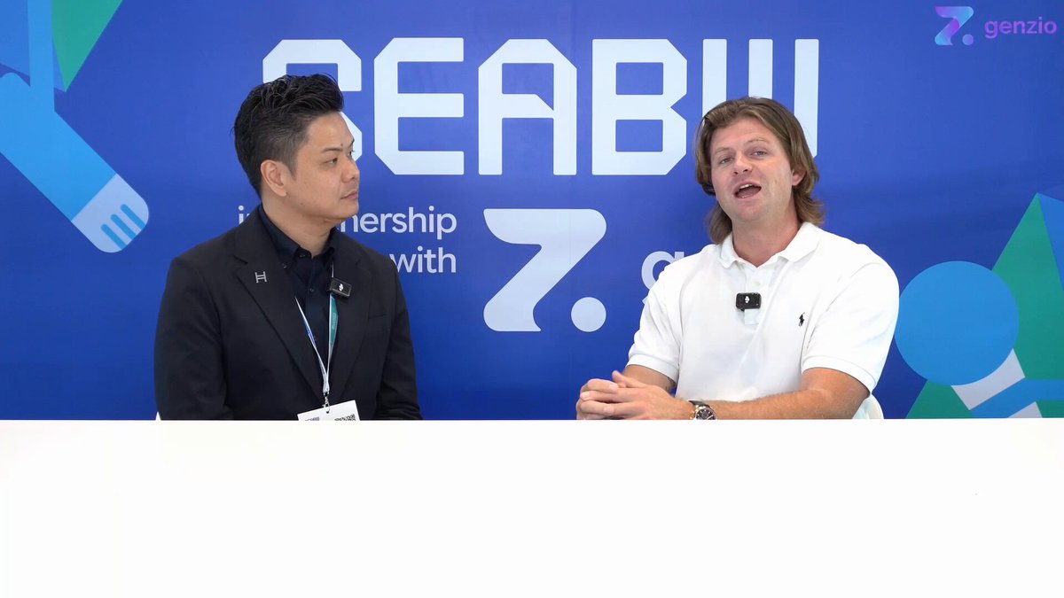 The image shows two men sitting across from each other at a table with a blue banner behind them that says "SEABWOfficial" and "@jtchui_abroad". One man has his arms crossed, while the other one is leaning forward in conversation. The background of the image features a logo for Genzio, which suggests they might be discussing opportunities emerging in Thailand or the role of real-world assets in driving adoption of cryptocurrencies.