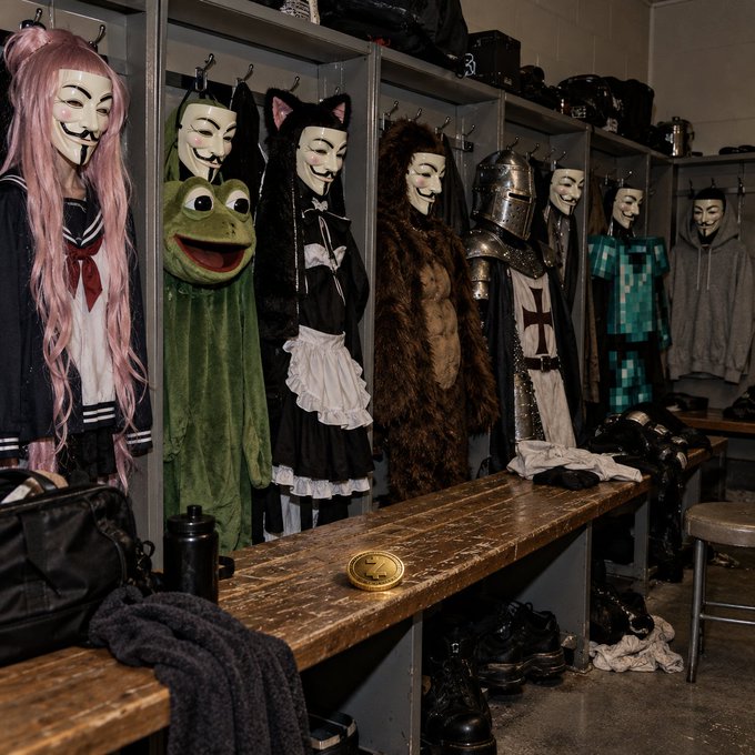 A locker room or backstage area displaying various costumes and Guy Fawkes masks hanging in cubbies, including outfits resembling a schoolgirl, a frog, a maid, a medieval soldier, and a creeper from Minecraft.