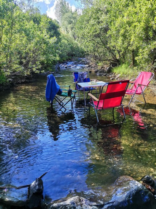 A colorful campsite is set up directly in the middle of a shallow stream, featuring pink and red folding chairs, a small table, and a blue towel hanging over a camping stool, surrounded by lush green trees.

The image contrasts sharply with the post's content: while the text celebrates financial success and discusses how to invest 10 billion VND, the photo depicts an escape from material pursuits—someone enjoying a simple, peaceful moment relaxing in nature rather than accumulating wealth.