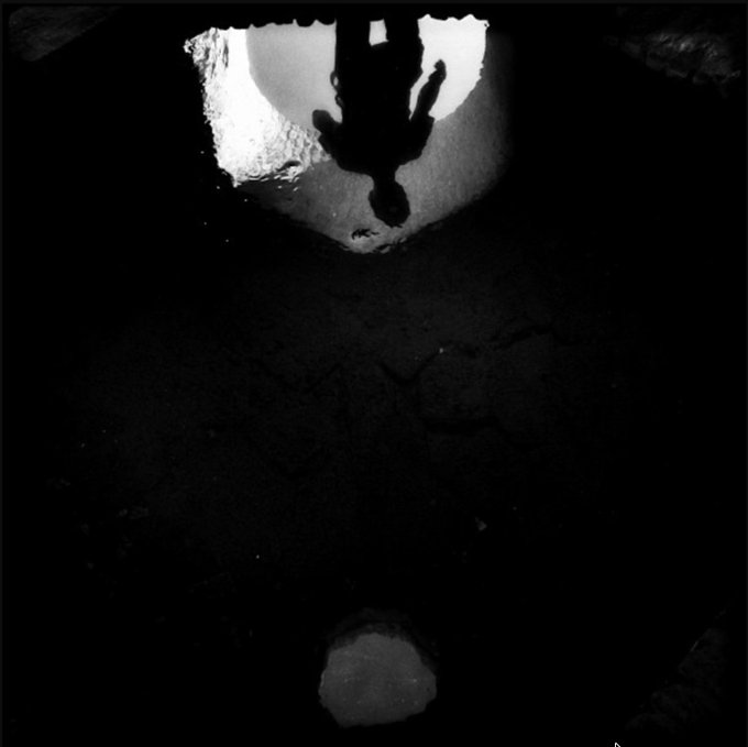 A dark, high-contrast black and white image showing a silhouette of a person reflected in a puddle on the ground.