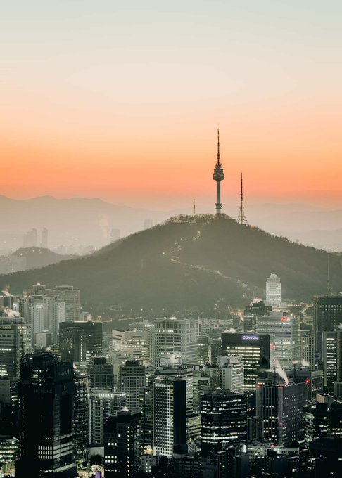 A view of Seoul's skyline at sunset, featuring the iconic Namsan Tower perched atop a mountain overlooking the dense cityscape below. The image visually anchors the caption's reflection on Seoul's rapid transformation, mirroring the linguistic shift in taxi drivers as a symbol of Korea's broader economic and cultural evolution.