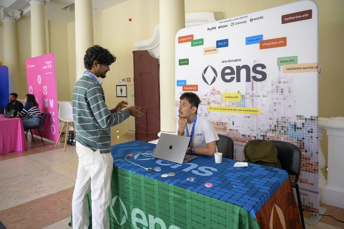 Two men converse at an ENS (Ethereum Name Service) booth at what appears to be a crypto or blockchain event, with the ENS branded tablecloth and backdrop displaying various .eth domains.