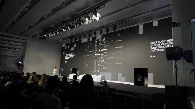 An indoor event photograph showing a presenter on stage in a dark conference room with a large screen displaying a website layout and the text 'BUILT TOGETHER. GLOBAL CONVER WITH OUR AFFIL PARTNERS.'