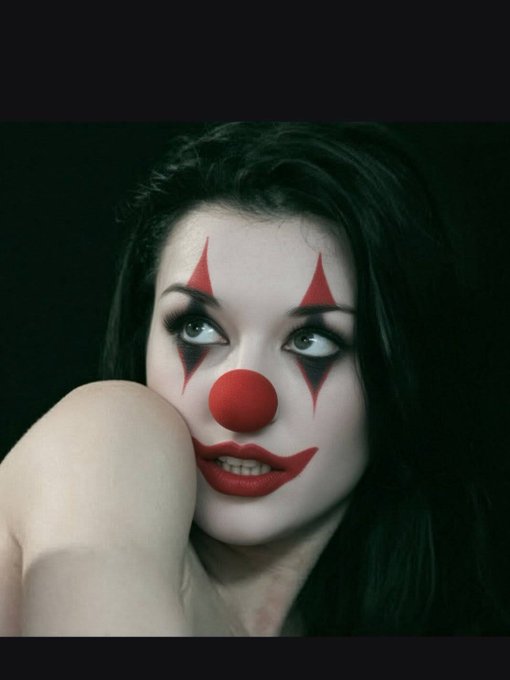 Dramatic portrait photograph of a woman with dark hair styled in elaborate clown makeup featuring red triangular designs around her eyes, a red clown nose, and red lips, photographed against a black background.