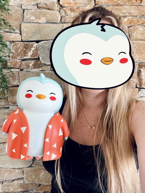 A blonde woman with her face covered by a cartoon penguin sticker, holding a matching penguin figurine wearing an orange kimono-style robe.