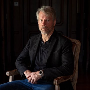 A portrait of Michael Saylor, chairman of MicroStrategy, seated in a dark wooden chair wearing a black blazer, accompanies news of his praise for the SEC's innovation exemption enabling 24/7 onchain trading of tokenized MSTR and STRC.