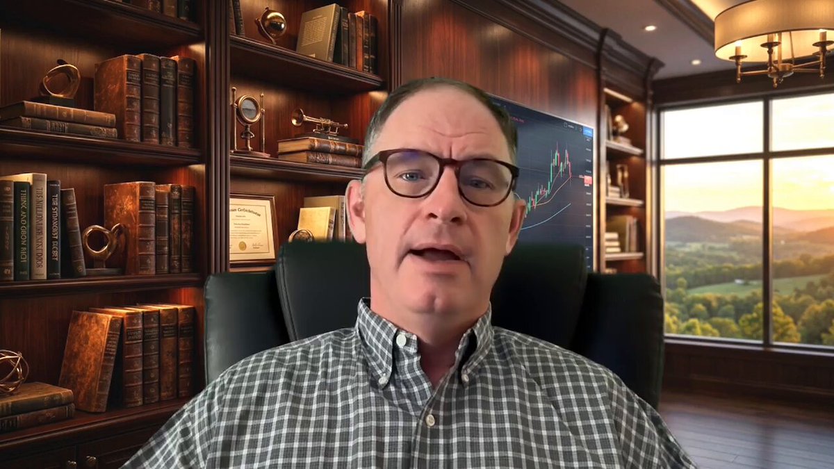 The image features a man sitting in an office chair, wearing glasses and a plaid shirt. He appears to be giving a presentation or speaking into a camera. The room has a window that offers a view of mountains outside, creating a scenic backdrop for the scene. There are several bookshelves filled with various books around the room, suggesting a well-read environment.