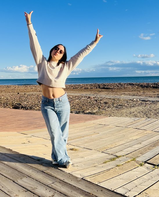 A woman in a cropped hoodie and jeans poses joyfully with arms raised on a wooden boardwalk by the seaside, capturing the uplifting, carefree energy of a "Gm fam" greeting with the sunny coastal backdrop.