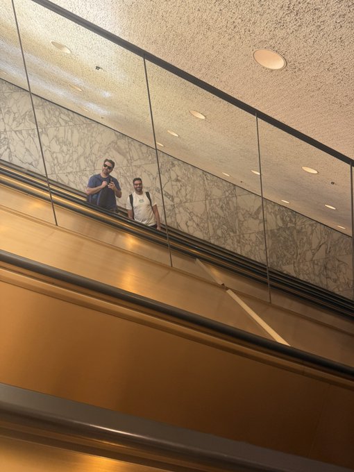 Two men stand on an upper level of a multi-story atrium with marble-patterned walls and glass railings, one holding a phone, looking down at the golden-toned lower floors.