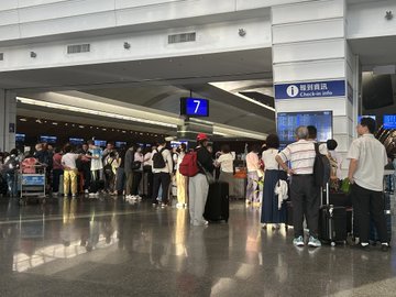 A crowd of travelers waits in line inside an airport terminal near gate 7, with an overhead sign reading '出行指引 Check-in guide' in Chinese and English.