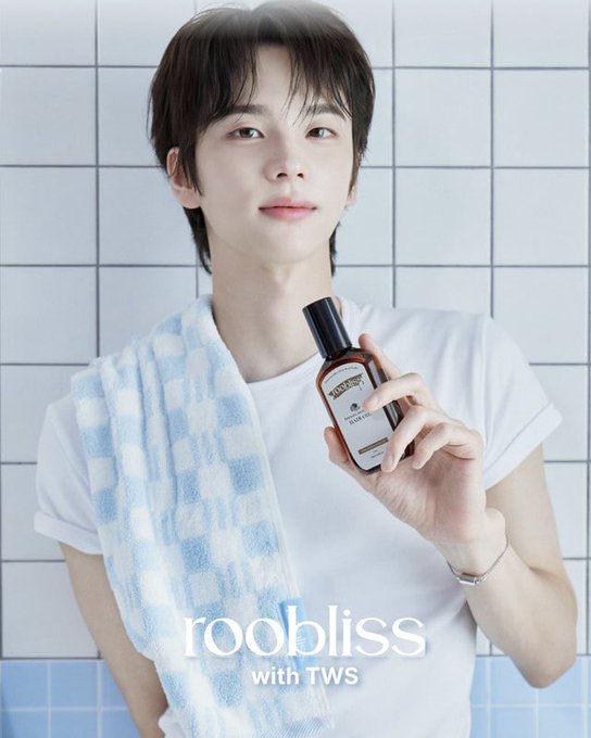 A young man with dark hair stands against a white tiled background, wearing a white t-shirt with a blue and white checkered towel draped over his shoulder while holding up a "roobliss" branded bottle. The text "roobliss with TWS" is overlaid at the bottom of the image, matching the caption identifying the subject as Shinyu in a promotional shoot for the brand's official website.