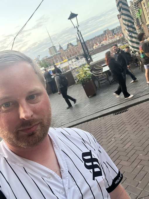 The image features a man wearing a baseball jersey with "S" written across his chest. He appears to be posing for a photo while standing near some potted plants. The background of the picture shows people walking around, indicating that they are likely in an outdoor setting or a public space like a park or a street. The post caption reads: "GM Have a great thursday, Don' forget to use Solflare". This suggests that the image was taken on Thursday and might have been posted on social media platforms for others to see.