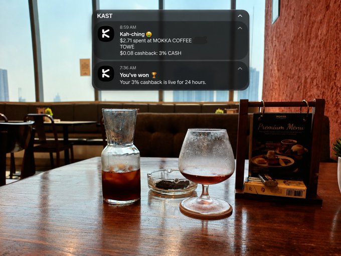 A coffee shop scene at a wooden table showing iced drinks and a 'Premium Menu' sign, with an overlaid KAST app notification showing a $2.71 cashback notification and a 3% cashback win at MOKKA COFFEE TOWIE.