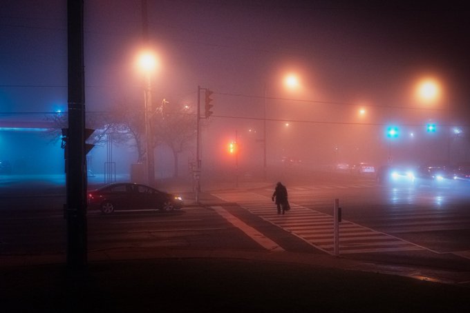 A foggy nighttime street intersection with glowing red and blue traffic lights, where a pedestrian is crossing the street.