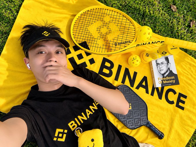 A selfie of a young man wearing a black Binance visor and t-shirt lying on a yellow Binance-branded mat on grass, surrounded by a yellow tennis racket, pickleball paddle, wiffle balls, and a book titled 'Freedom of Money' featuring CZ's portrait.