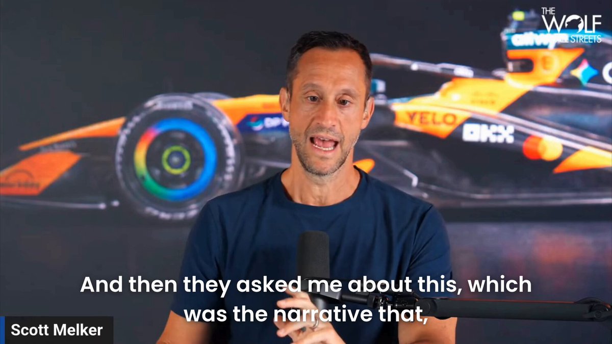 The image features a man wearing a blue shirt holding a microphone, talking into it as if he's giving an interview or sharing his thoughts about Bitcoin. The background of the image shows a car with a tire visible behind him, suggesting that they are in a setting related to the automobile industry.