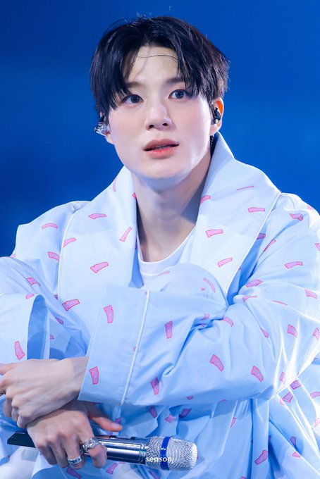 A young man with dark hair wears a light blue oversized jacket with a pink pattern and holds a microphone during a performance, with a "SHOPOIL" watermark visible. The image appears to be a concert photo likely referenced in the linked X (Twitter) post.