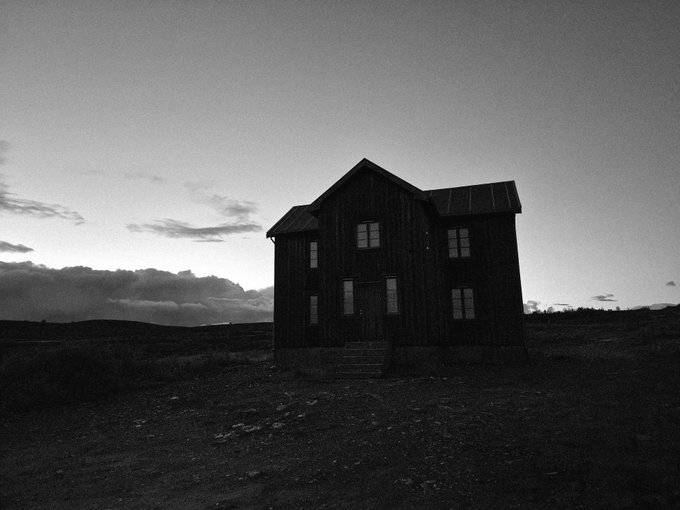A black and white photograph of a dark two-story house with solar panels on the roof, silhouetted against a cloudy sky in a barren landscape at dusk.